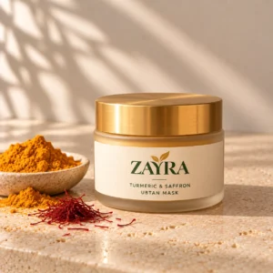 ZAYRA Turmeric & Saffron Ubtan Mask in a frosted glass jar with a brushed gold lid, beside turmeric powder and saffron threads